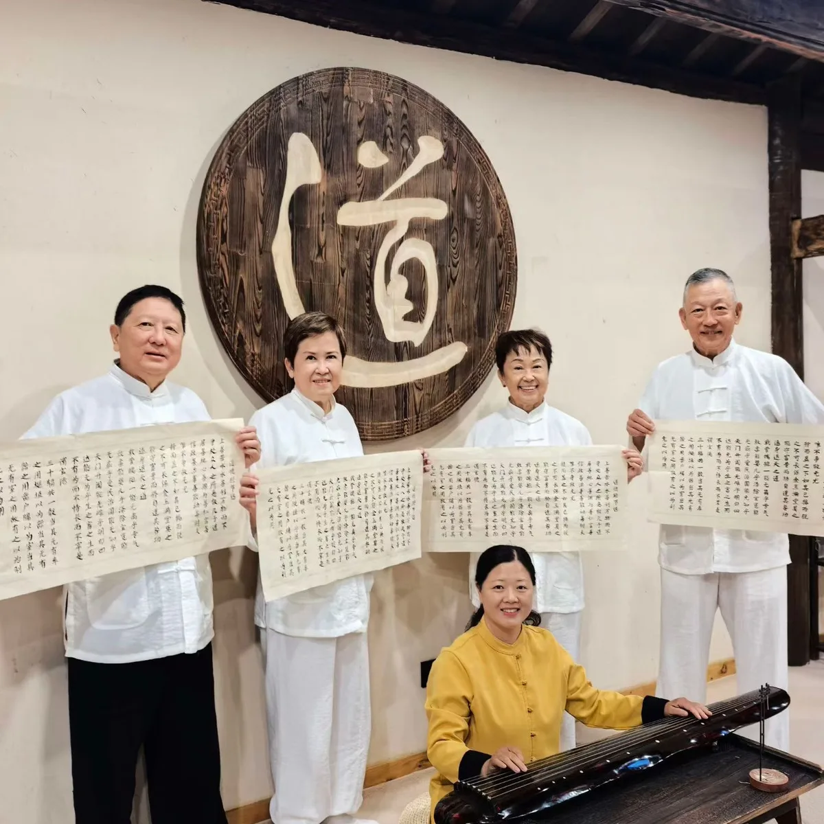 Quatre participants présentent leurs rouleaux de calligraphie sous une plaque portant le caractère Dao, avec un guqin au premier plan.