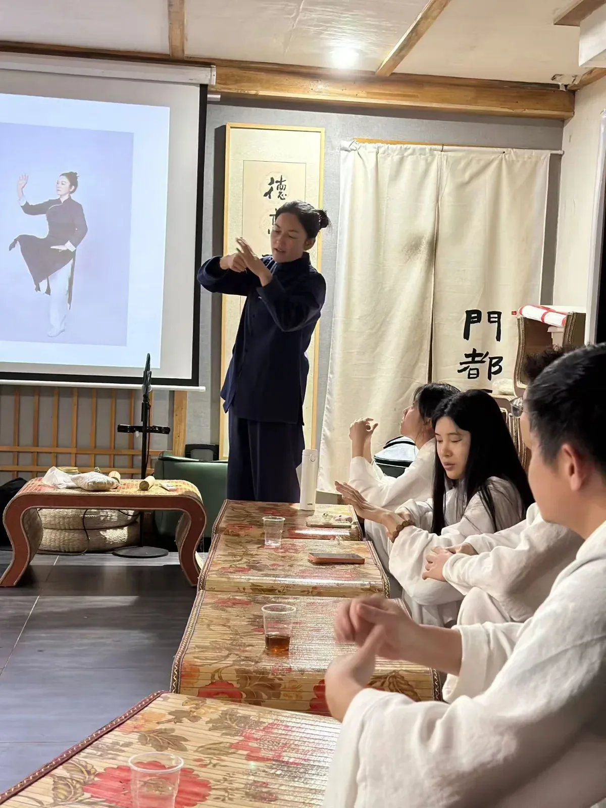 Un enseignant en bleu sombre montre une position de la main devant une photographie projetée de tai-chi ; les élèves reproduisent le geste.
