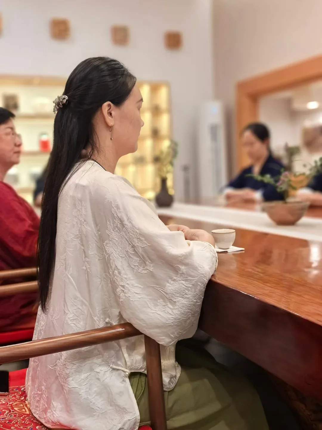 A woman in an embroidered white silk jacket sits upright at a polished wooden table, hands resting beside a small white cup, waiting.