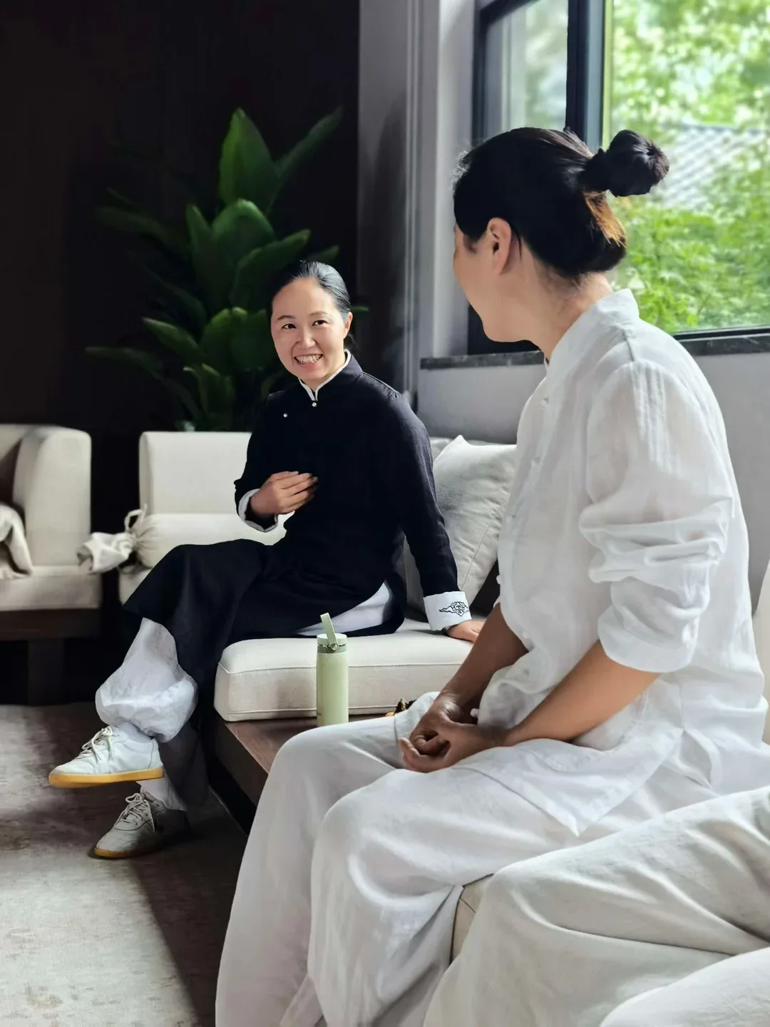 Deux femmes conversent sur un canapé clair près d’une fenêtre ; celle en robe noire sourit en parlant, tandis que celle en blanc écoute.