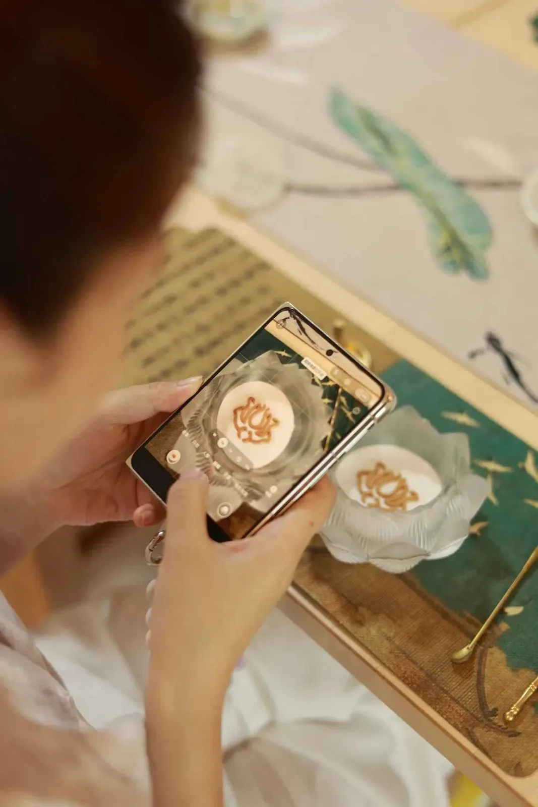 Une personne photographie le sceau d’encens terminé ; le brûle-parfum repose à côté du téléphone et des outils sur un tapis bleu-vert.