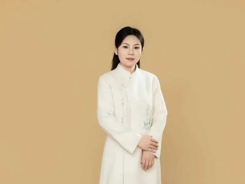 Chongmin en robe blanche brodée, les mains croisées, devant un fond uni beige.