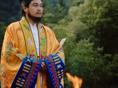 Teacher Facheng in ceremonial Daoist ritual vestments, holding a document, beside a fire in a forest clearing.