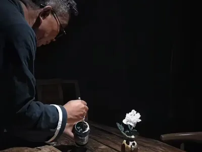 Chen Haichao leans over a wooden studio table by lamplight, brush in hand beside an ink jar and a white peony.