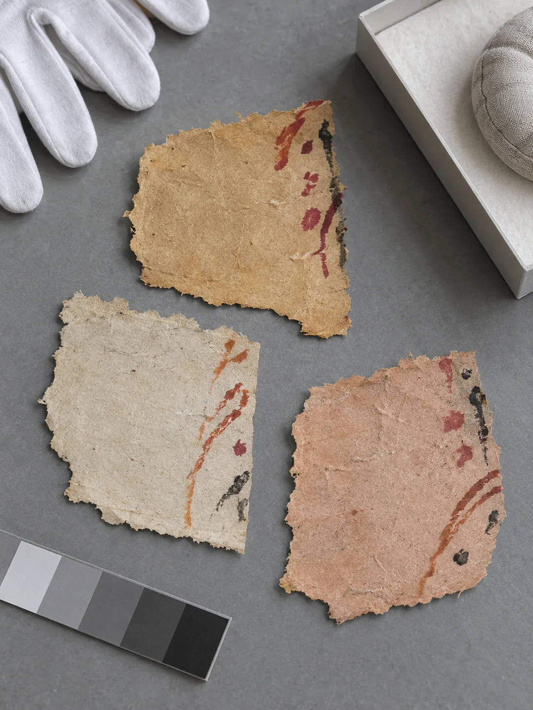 Photographie d’étude générée par IA, vue du dessus, de trois fragments fictifs de papier fibreux portant des marques abstraites en bordure sur une table de conservation gris froid
